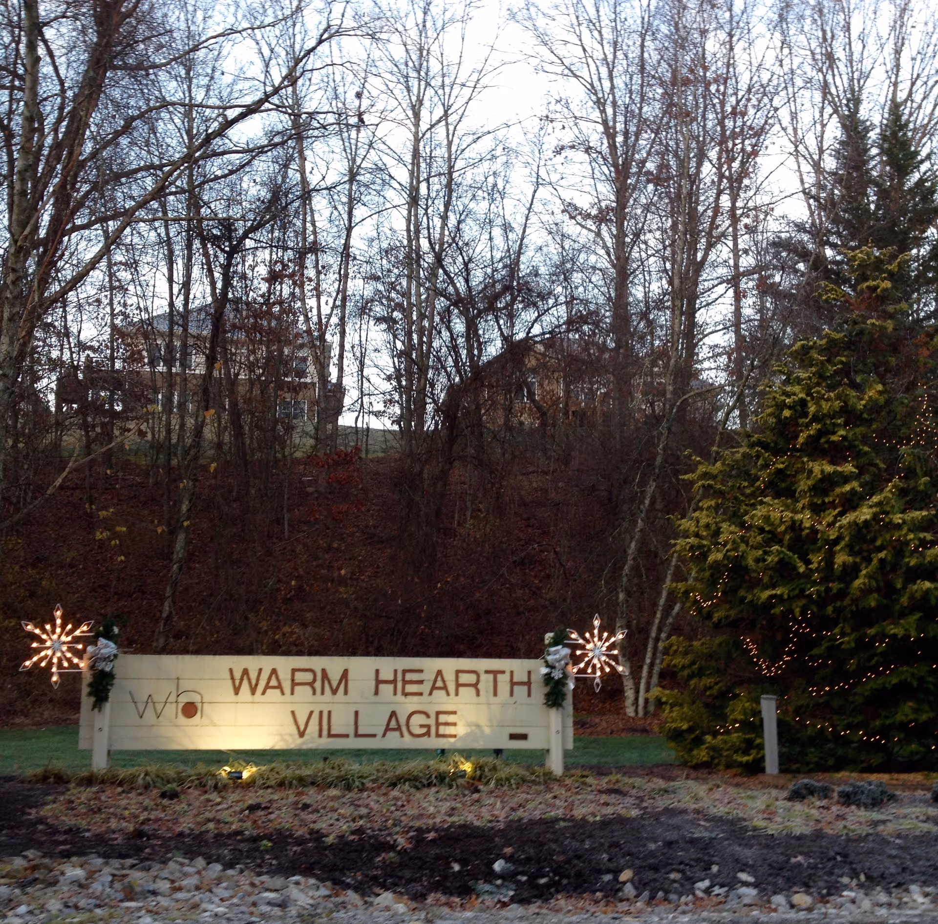 Outdoor view of a sign that reads 'Warm Hearth Village' with decorative lights and greenery around it. Behind the sign are leafless trees and a couple of houses on a hill.