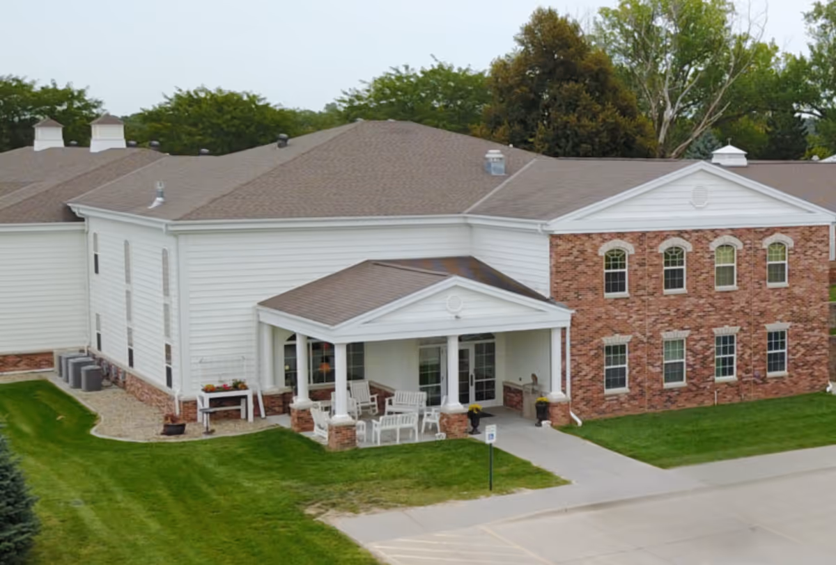 Exterior view of a senior living facility building with white siding and red brick accents. The building has a covered entrance with white columns and outdoor seating including chairs and a bench. There is a well-maintained lawn and a paved driveway in front.