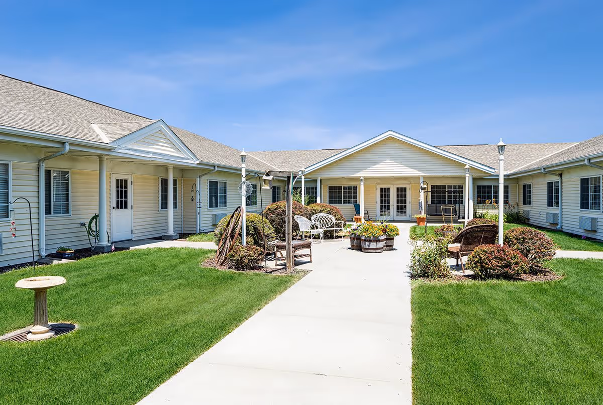 Outdoor courtyard area of Morton Memory Care facility with green grass, bushes, patio furniture, and a concrete walkway leading to a building with white siding and multiple windows under a clear blue sky.