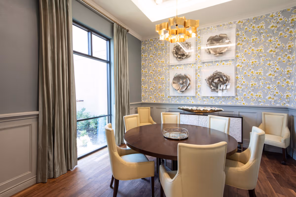 A dining room with a round wooden table surrounded by eight beige upholstered chairs. The room features floral wallpaper with framed 3D flower art pieces on the wall, a modern gold chandelier, large window with beige curtains, and wooden flooring.