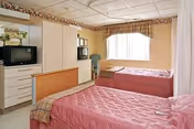 Two-bed resident room with pink quilted beds, a dresser holding a television, a window with valances, and a privacy curtain.