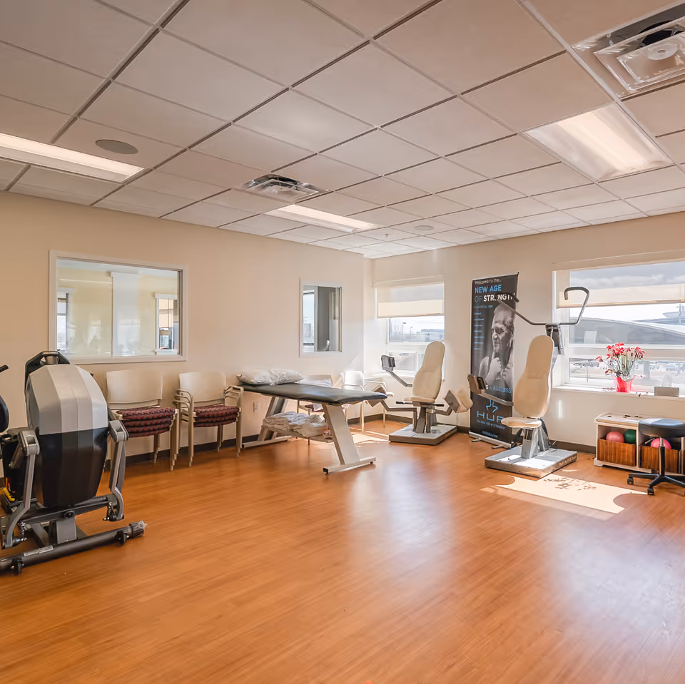 Sunlit rehabilitation/exercise room with therapy chairs, a treatment table, exercise equipment, and stacked visitor chairs.