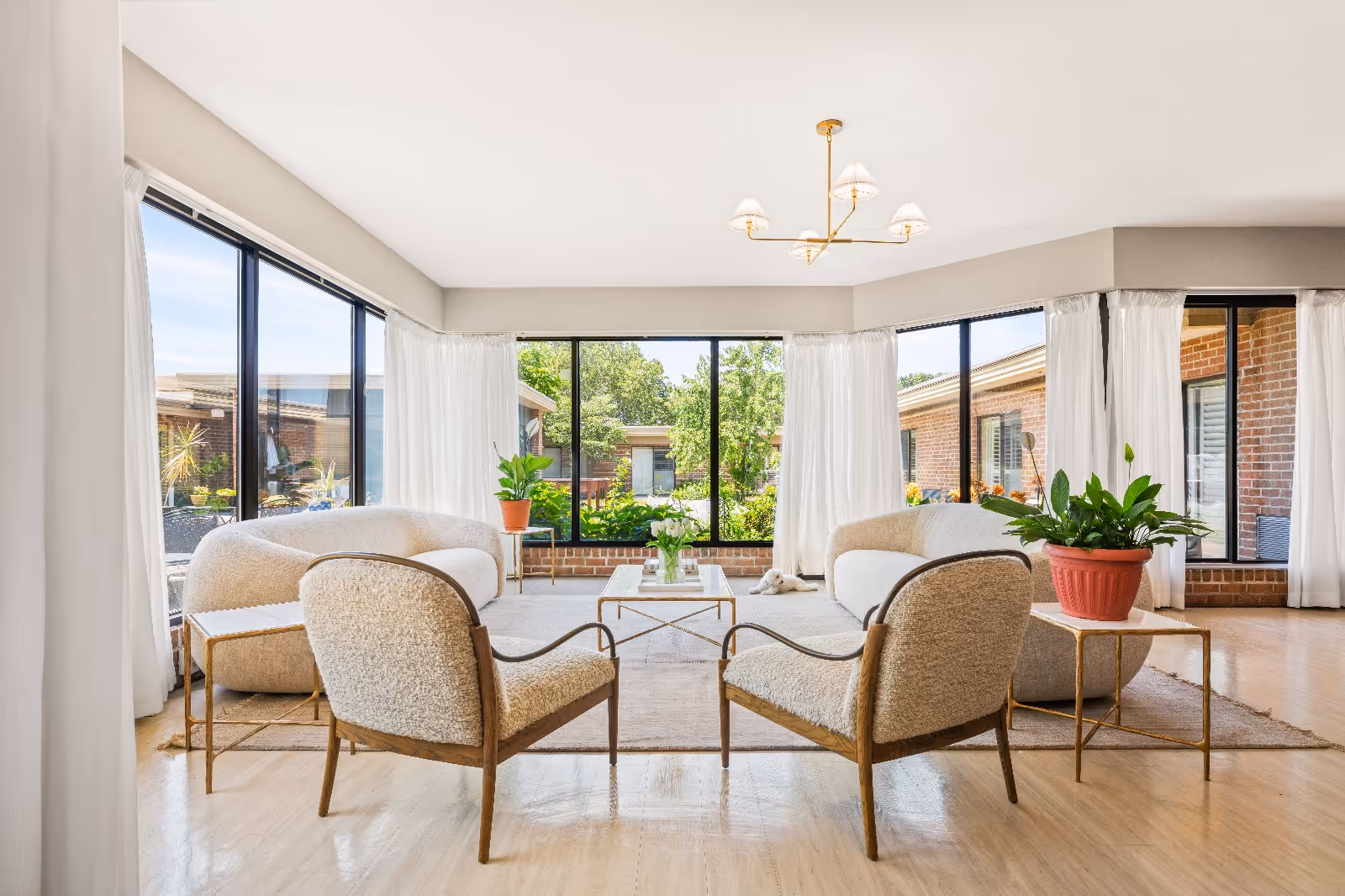 Bright and airy living room with large windows showing greenery outside. The room features two beige armchairs and two matching sofas arranged around a glass coffee table on a light-colored rug. There are potted plants on side tables and a modern chandelier hanging from the ceiling.