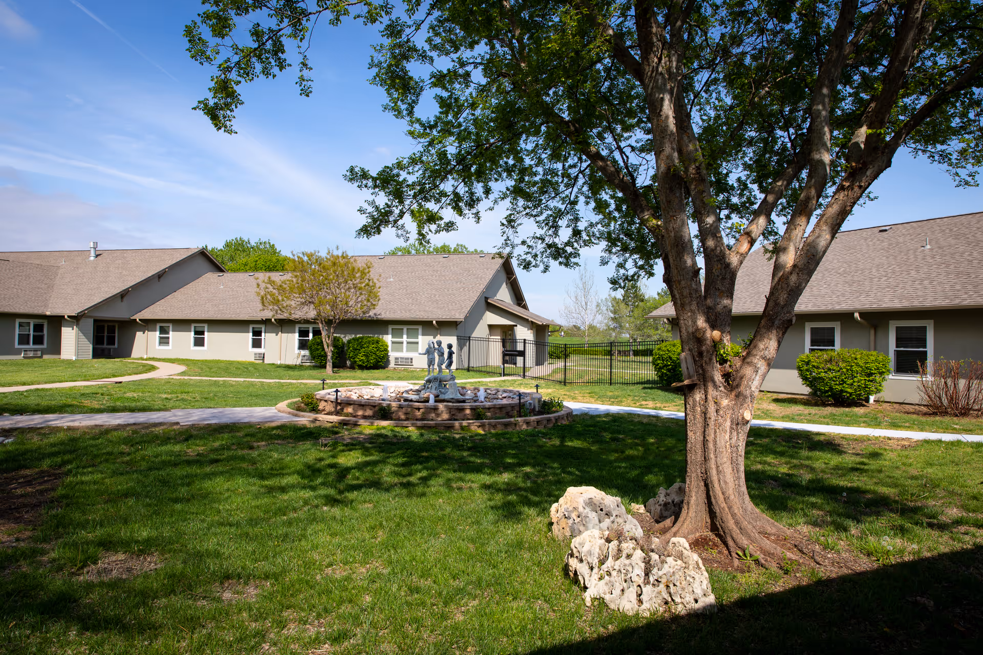 Courtyard with grass, a large tree, a central fountain sculpture, and surrounding single-story assisted living buildings under a blue sky.