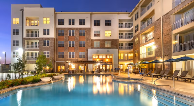Evening view of a modern multi-story residential building with balconies overlooking a large outdoor swimming pool surrounded by lounge chairs and umbrellas, with lights reflecting on the water.