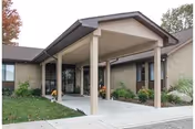 Entrance of Belmont Healthcare Center showing a covered drop-off area with a peaked roof supported by columns, surrounded by a brick building and landscaped greenery.