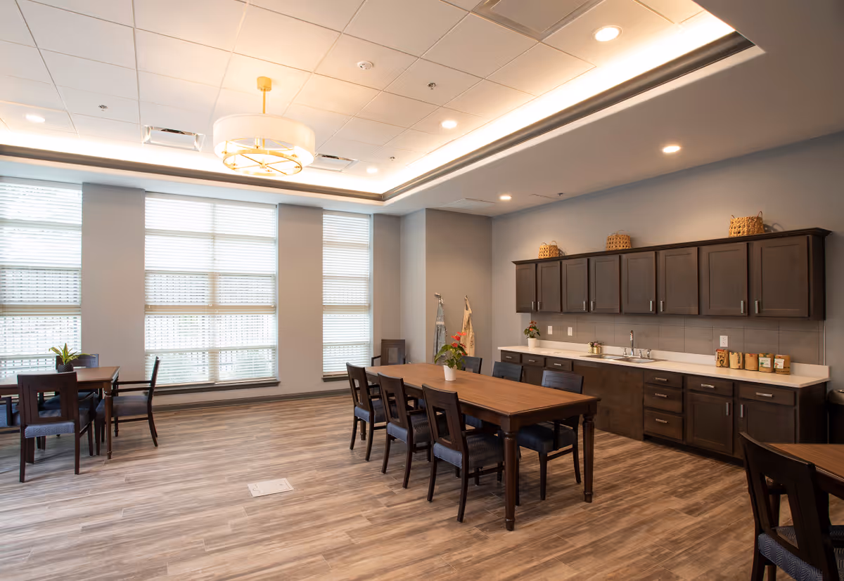 A spacious, well-lit dining area with large windows covered by blinds, wooden tables and chairs, and a long counter with dark wood cabinets and a sink. The ceiling features recessed lighting and a modern chandelier.