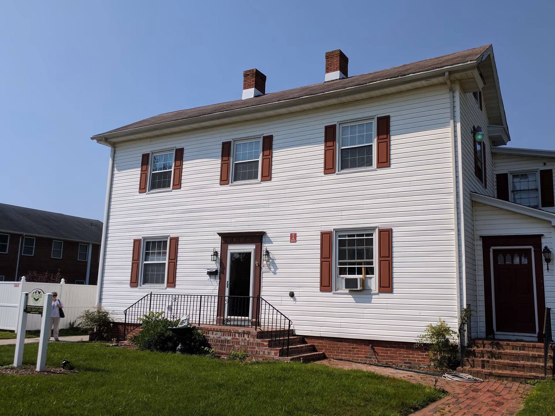Front view of a two-story white vinyl-sided house with red shutters, brick steps, a lawn and a sign in the yard.