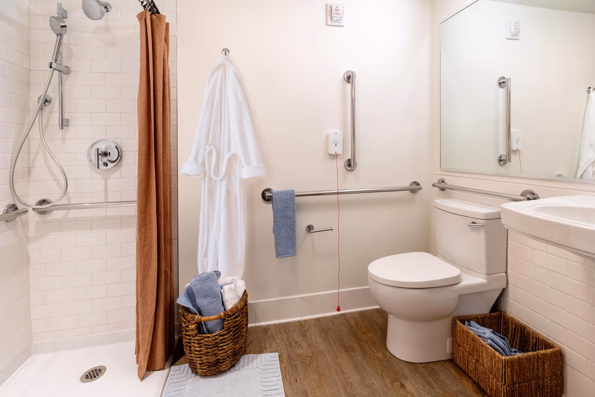 A clean and accessible bathroom featuring a walk-in shower with a brown curtain, white tiled walls, and grab bars. A white bathrobe hangs on the wall next to a basket filled with folded towels. The bathroom also includes a toilet with grab bars, a sink with a large mirror above it, and a small basket with towels on the floor. The floor is wood-style, and there is an emergency pull cord on the wall.