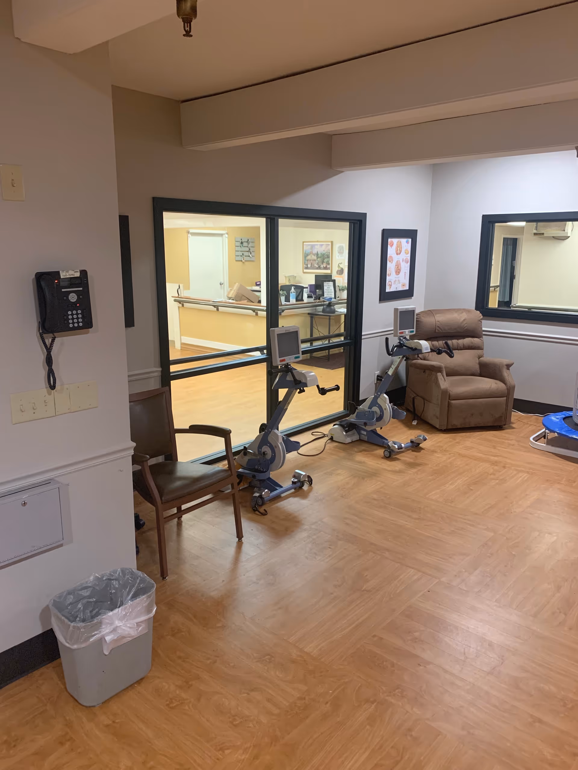 An interior room with two exercise bikes, a brown recliner chair, a wooden chair, and a small blue trampoline. The room has wood-patterned flooring and light-colored walls. There is a wall-mounted phone on the left and a large window looking into another room with a reception desk and chairs.