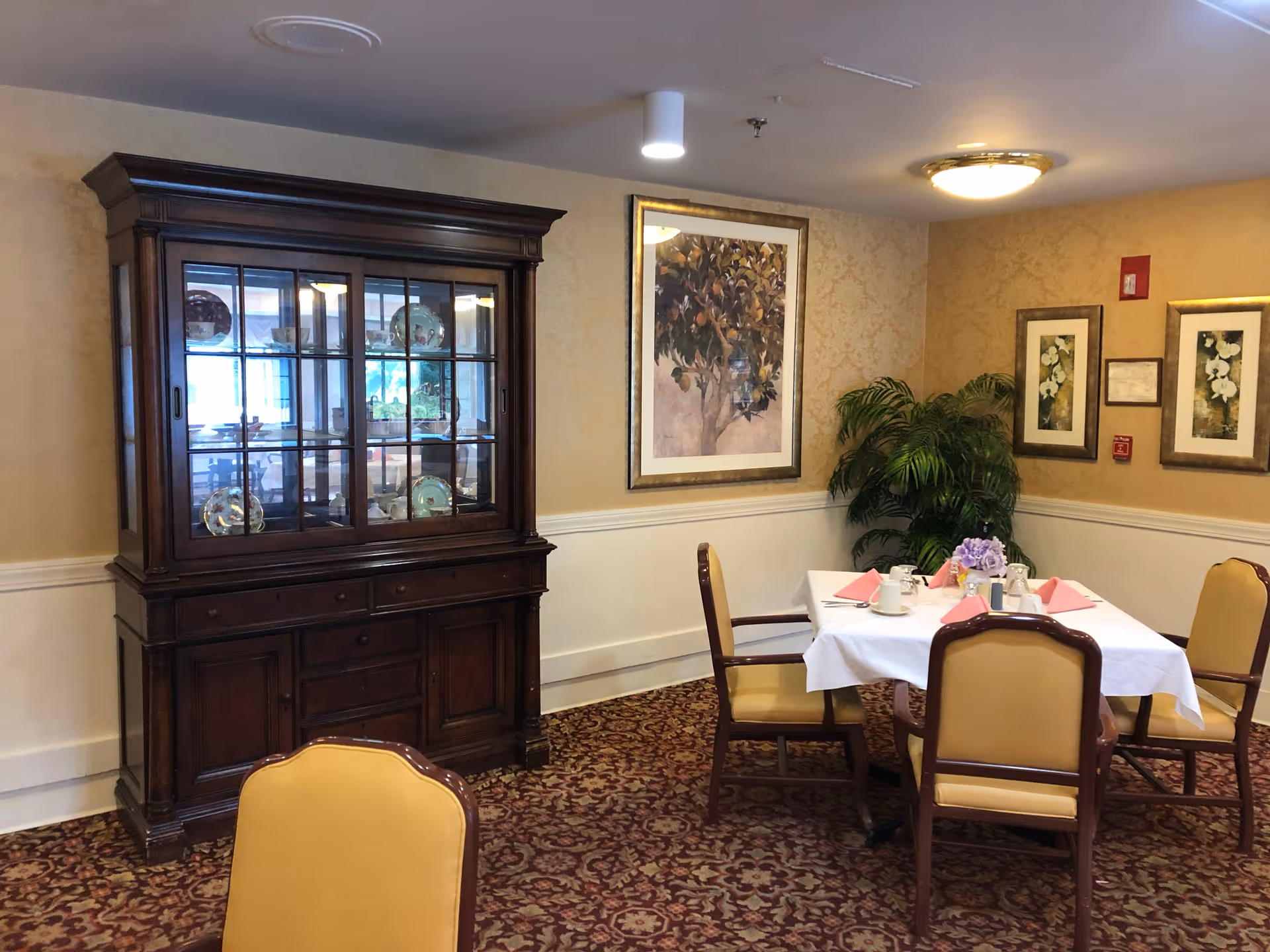 A dining area in a senior living facility with a wooden china cabinet displaying decorative plates, a table set with a white tablecloth, pink napkins, cups, and a floral centerpiece. The room has patterned carpet, beige wallpaper with framed artwork, and a potted plant in the corner.