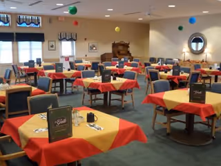 A dining room with multiple tables covered in yellow and red tablecloths. Each table is set with menus, cups, and silverware. The room has blue cushioned chairs, carpeted floor, and neutral-colored walls. There are colorful hanging decorations from the ceiling and framed pictures on the walls.
