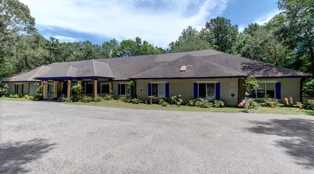 Single-story building with a dark shingled roof and beige exterior walls, surrounded by greenery and trees under a partly cloudy sky. The building has several windows with blue shutters and a covered entrance with columns. There is a paved area in front of the building.