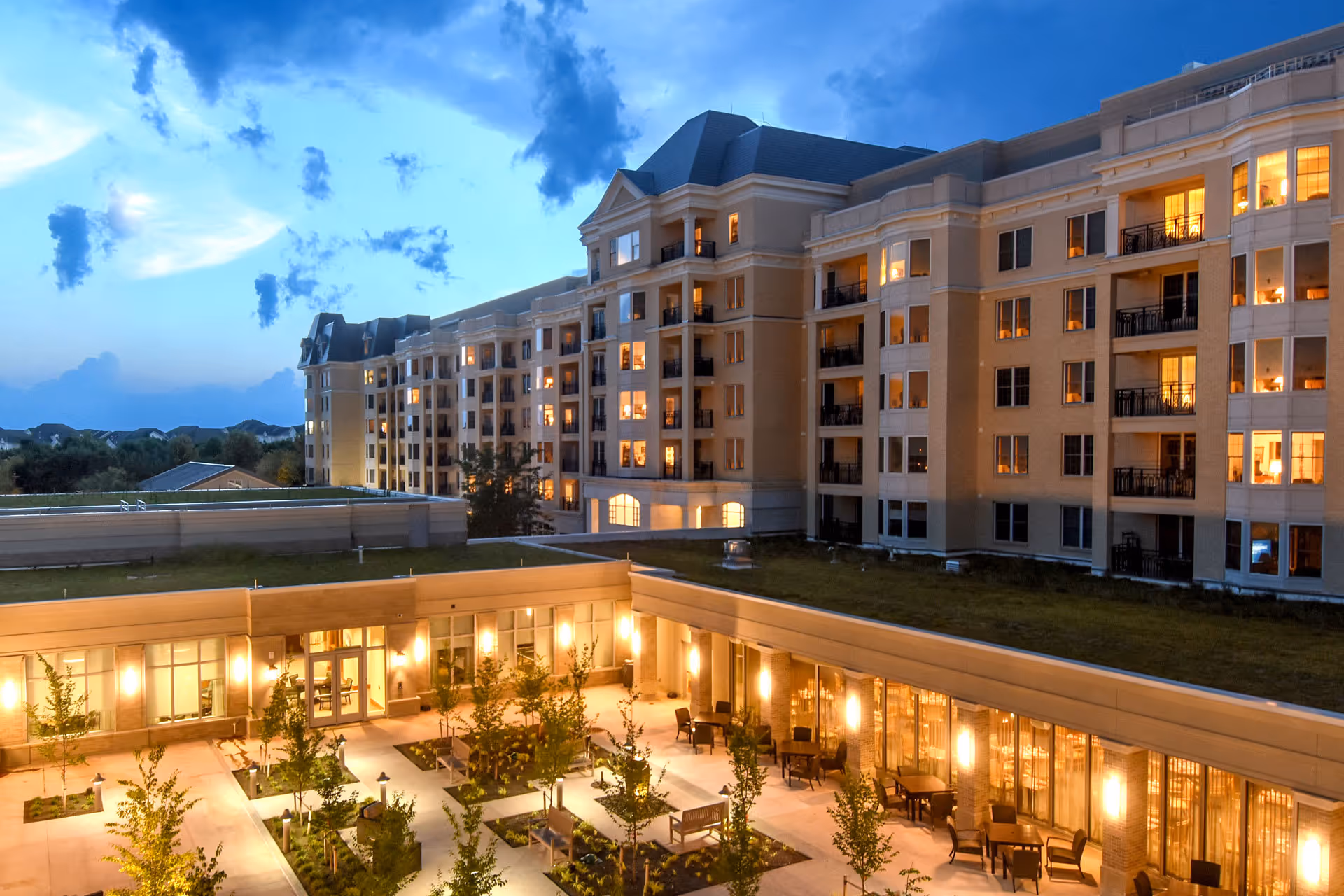 Evening view of Ingleside at King Farm senior living facility showing a large multi-story building with many lit windows and balconies surrounding a well-lit courtyard with benches, tables, chairs, and small trees.