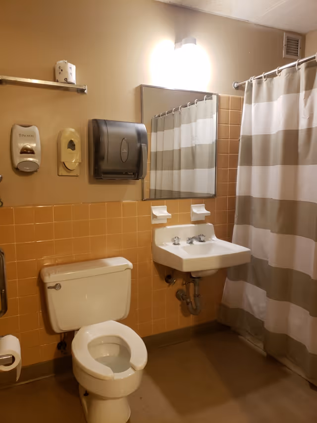 A bathroom with beige and tan tiles on the walls, a white toilet with an open seat, a white sink with two faucets, a large mirror above the sink, a striped beige and white shower curtain, a wall-mounted soap dispenser, a paper towel dispenser, and a toilet paper holder with a roll of toilet paper.
