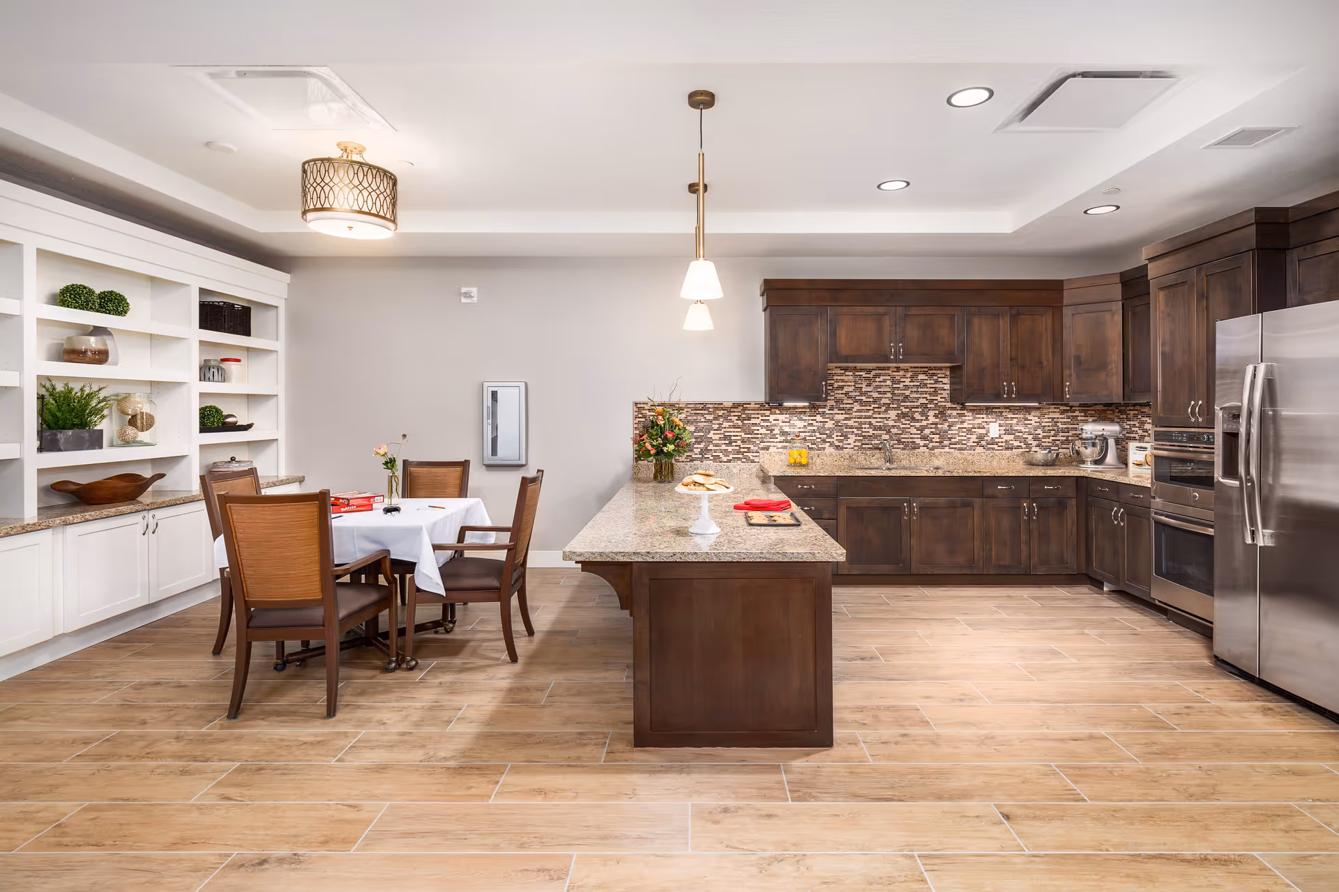 A spacious kitchen and dining area featuring dark wooden cabinets, a large kitchen island with granite countertop, stainless steel appliances including a refrigerator and oven, and a dining table with four chairs covered with a white tablecloth. The room has light wood flooring, built-in white shelves with decorative items, and modern ceiling lights.