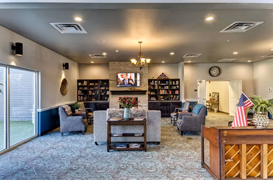 Spacious assisted-living common room with sofas and armchairs facing a TV above a stone fireplace, bookshelves, chandelier, and a piano with American flags.