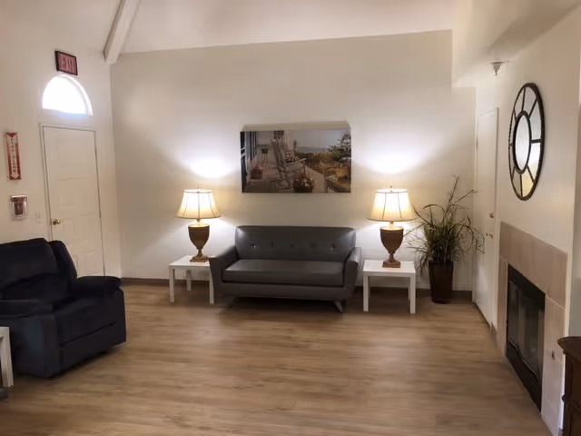 A cozy living room area with a gray loveseat centered between two white side tables, each holding a lamp with a beige shade. To the left is a dark recliner chair. On the wall above the loveseat is a framed picture of a porch with rocking chairs and a scenic view. To the right is a tall plant in a dark pot and a large round wall clock above a fireplace with a light-colored mantel. The floor is wood, and the walls are painted white. A door with an arched window above it is visible on the left side.