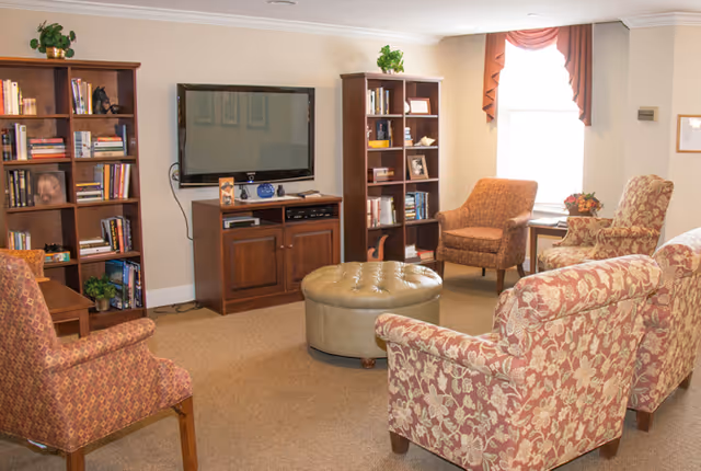 A cozy living room with patterned armchairs and a round tufted ottoman in the center. There are two wooden bookshelves filled with books and decorative items, and a flat-screen TV mounted on the wall above a wooden cabinet. A window with a valance allows natural light into the room.