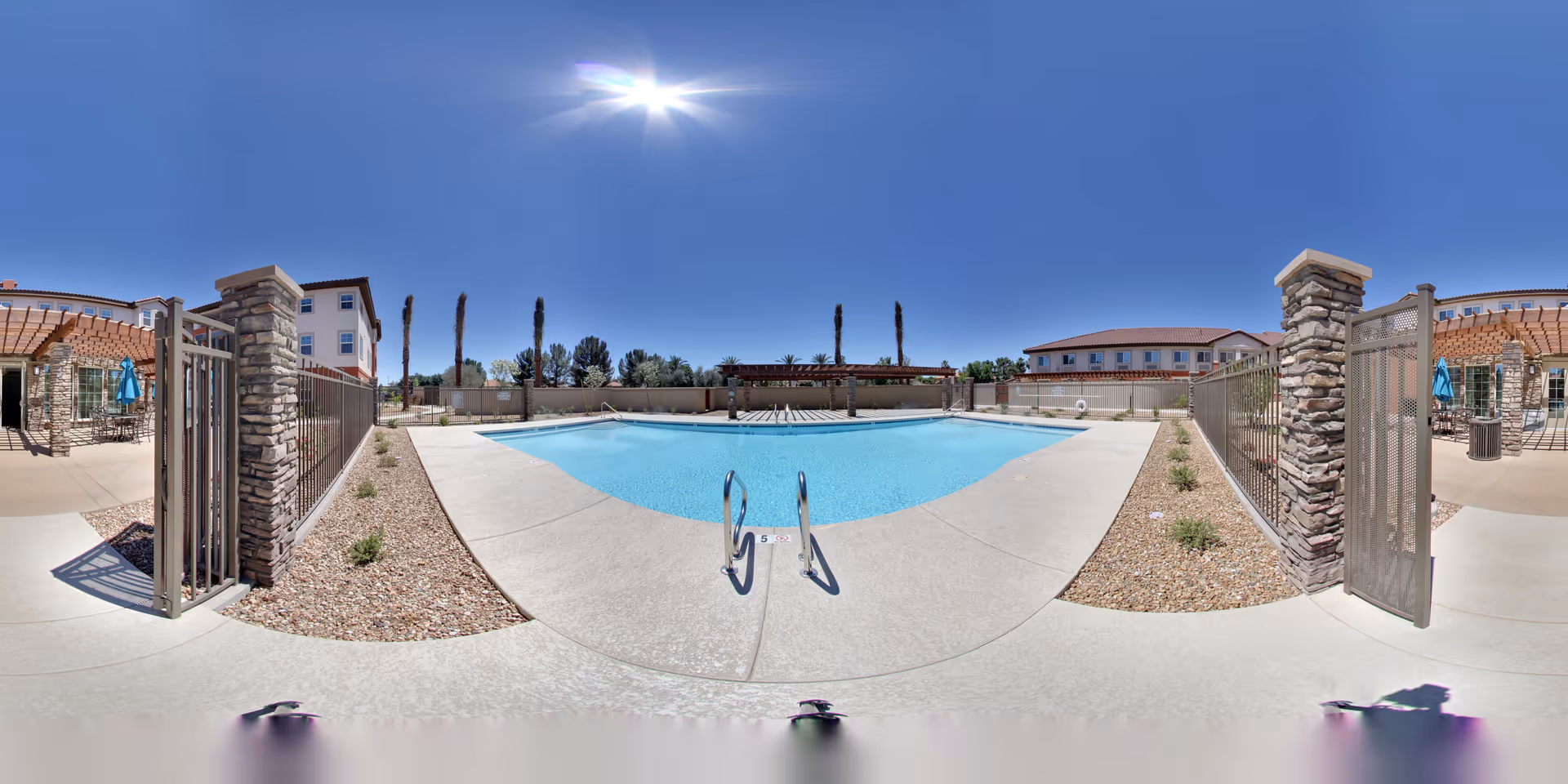 Sunlit outdoor swimming pool with gated entry, lounge seating and surrounding senior living buildings under a clear blue sky.