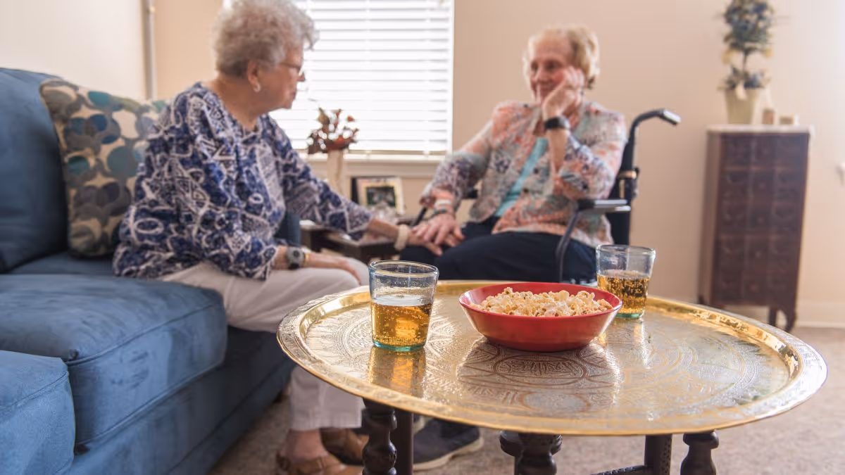 Two elderly women sitting in a living room, one on a blue couch and the other in a wheelchair, holding hands and engaging in conversation. In the foreground, there is a round brass tray table with a red bowl of popcorn and two glasses of a light-colored beverage. The room has a window with blinds, a small cabinet with a plant, and framed photos on a side table.