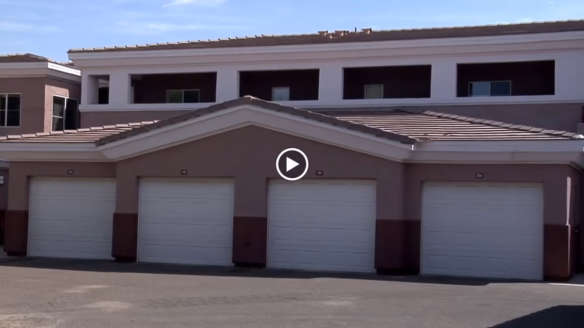 Exterior view of Chaparral Winds Assisted Living showing a row of four closed white garage doors beneath a two-story building with beige and brown walls and a tiled roof.