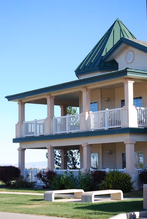 Two-story building with a green pointed roof and beige exterior, featuring spacious balconies with white railings on both floors. The building is surrounded by green grass, bushes, and small trees under a clear blue sky.