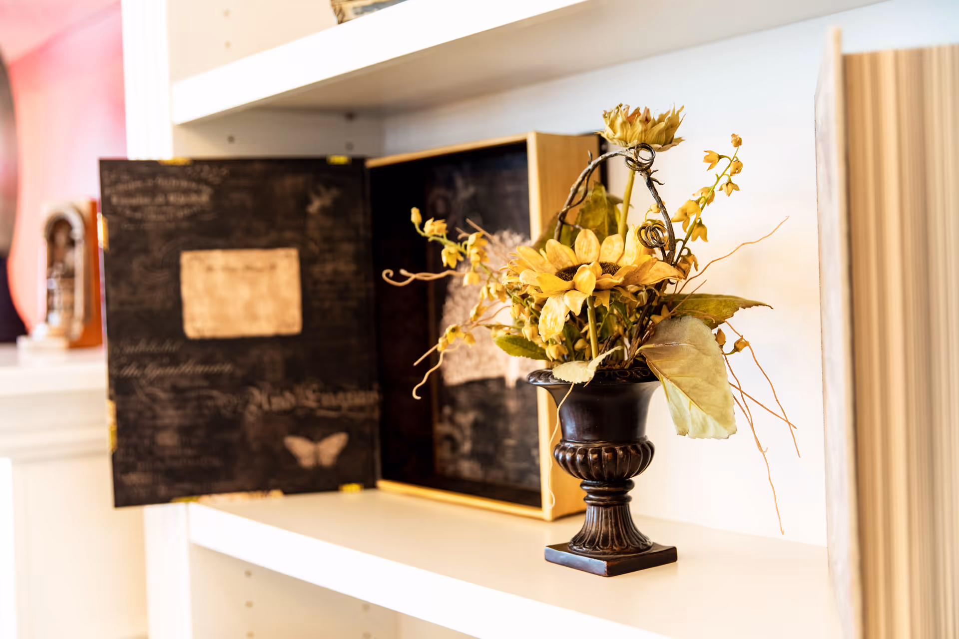 A decorative black vase with artificial yellow flowers and green leaves placed on a white shelf. Behind the vase, there is an open book with dark pages featuring faded text and a butterfly illustration. The background includes a blurred view of a room with a clock on a mantel.