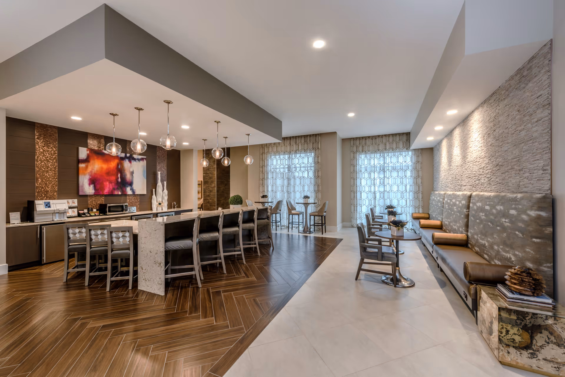 A modern and stylish common area with a long kitchen island featuring bar stools, pendant lights hanging above, and a countertop with a microwave and coffee machine. The space has a combination of wood and tile flooring, with seating areas including small tables and chairs near large windows with patterned curtains. The right side features a cushioned bench with pillows against a textured wall, illuminated by recessed lighting.