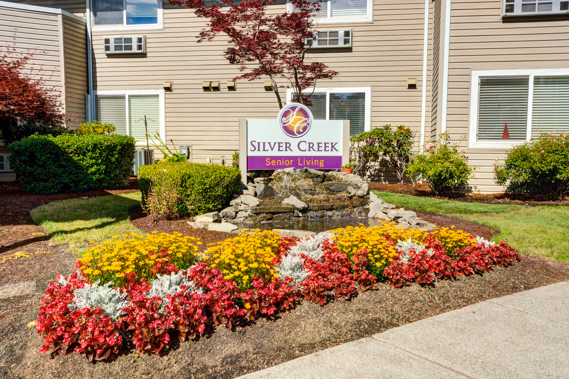 Front exterior view of Silver Creek Senior Living facility with a landscaped garden featuring colorful flowers, bushes, a small rock water fountain, and the facility's sign in front of a beige building with windows.