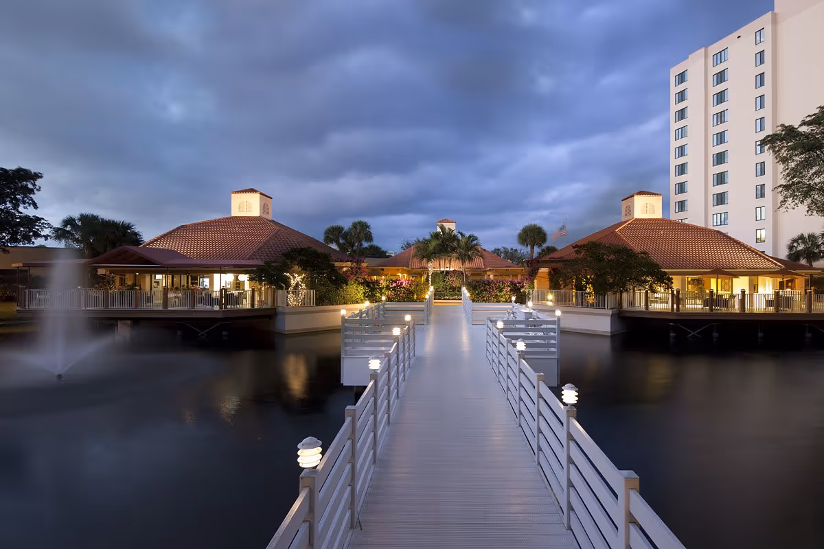 Evening view of The Waterford facility showing a well-lit walkway over water leading to buildings with red-tiled roofs surrounded by palm trees and landscaping, with a tall building in the background and a water fountain on the left side.
