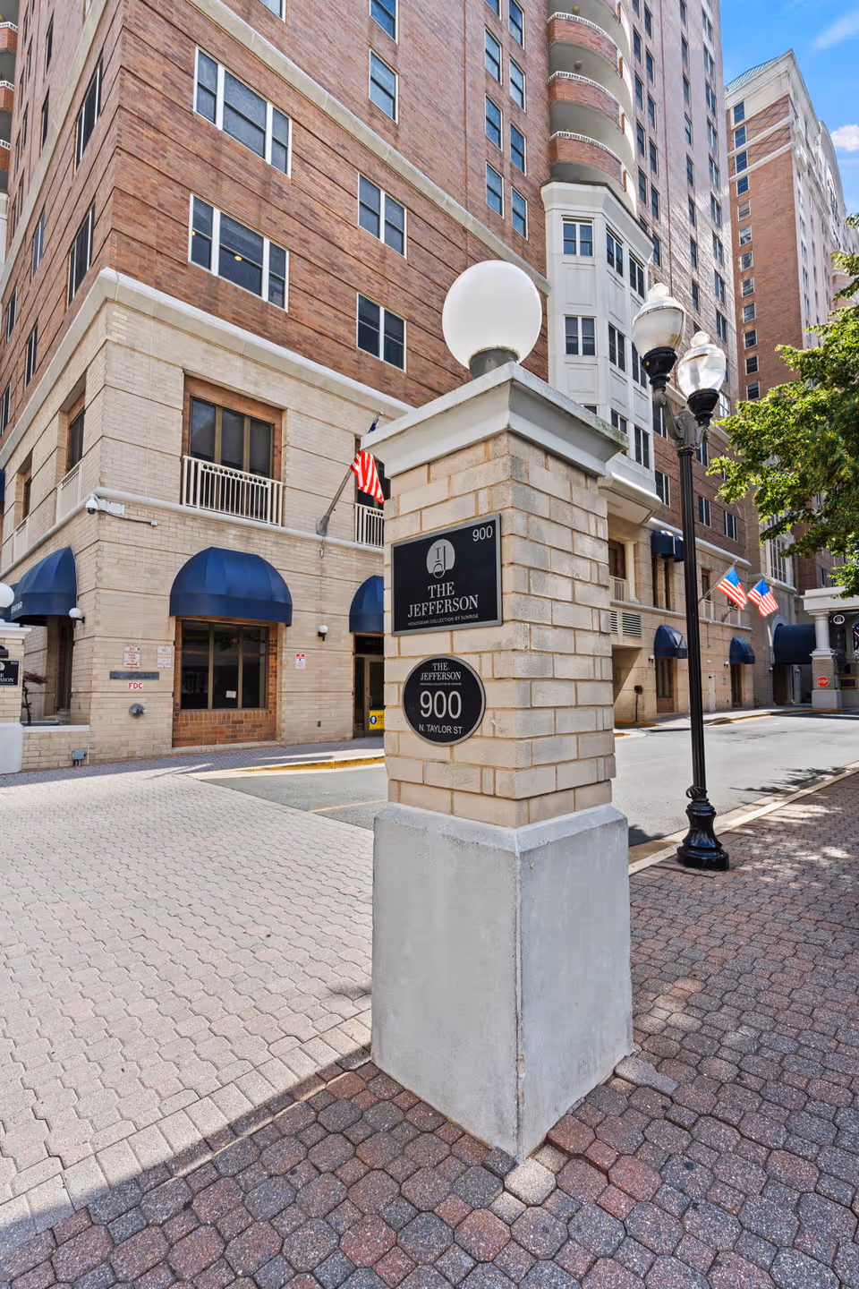 Exterior view of a multi-story brick building with blue awnings over the windows and entrances. A stone pillar in the foreground displays signs for The Jefferson at 900 N. Taylor St. There are street lamps and American flags along the sidewalk under a blue sky with some clouds.