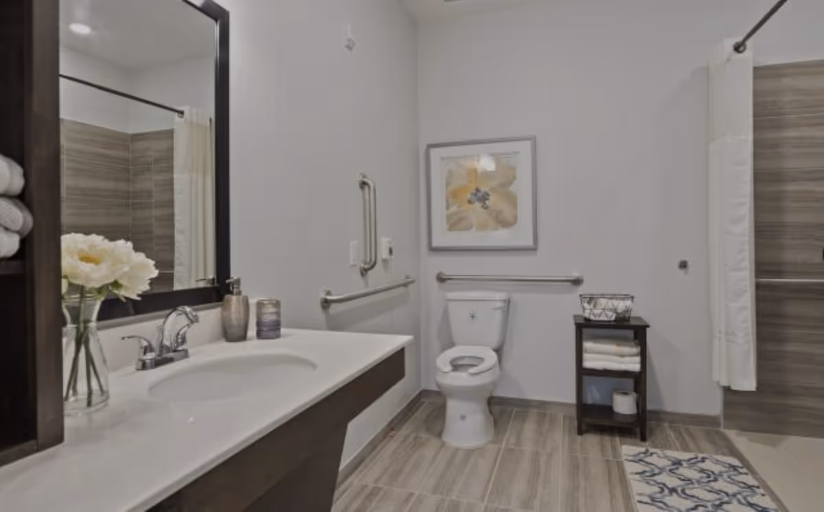 A clean and modern bathroom featuring a white sink with a silver faucet, a large mirror, a vase with white flowers, a toilet with grab bars on the walls, a small shelf holding towels and toilet paper, and a shower area with a curtain and tiled walls.