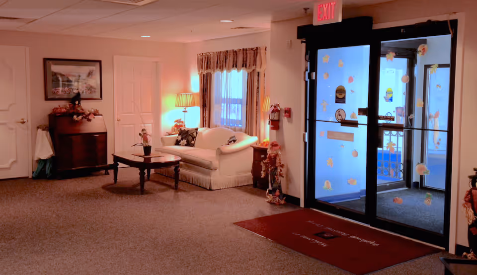 Interior view of a senior living facility lobby area with a white couch, wooden coffee table, side tables with lamps, a framed picture on the wall, and glass entrance doors decorated with autumn-themed stickers. There is a red welcome mat on the floor and an exit sign above the door.