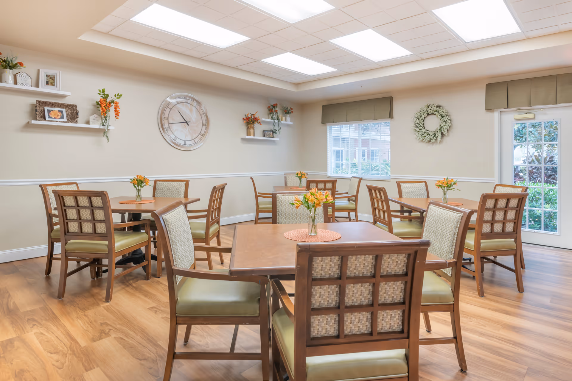 Bright communal dining room with wooden tables and chairs, small floral centerpieces, wall clock, and windows.