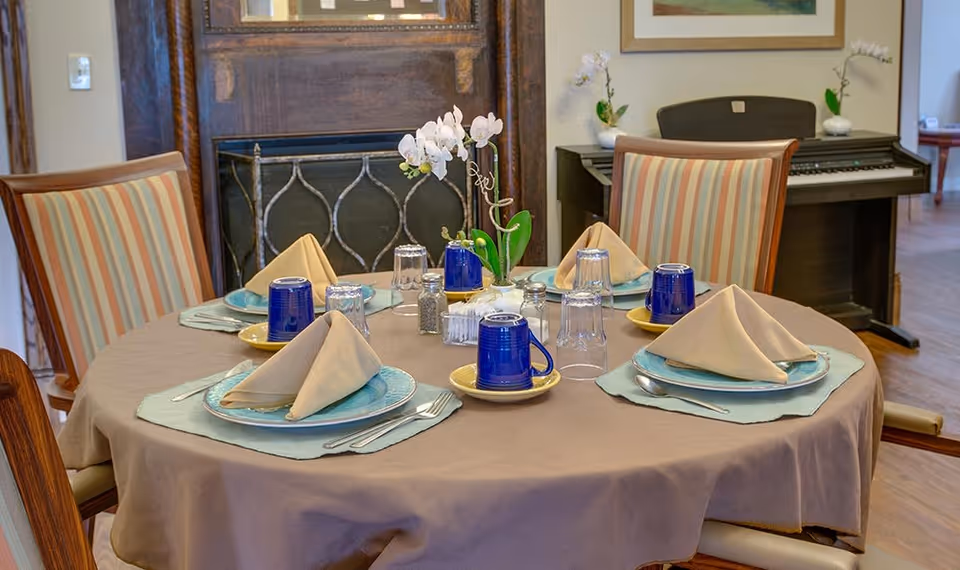 A round dining table set for four with beige napkins folded on blue plates, blue cups on yellow saucers, silverware, and a small white orchid centerpiece. The table is covered with a brown tablecloth and light blue placemats. In the background, there is a piano with two small white orchid plants on top and a framed picture on the wall.