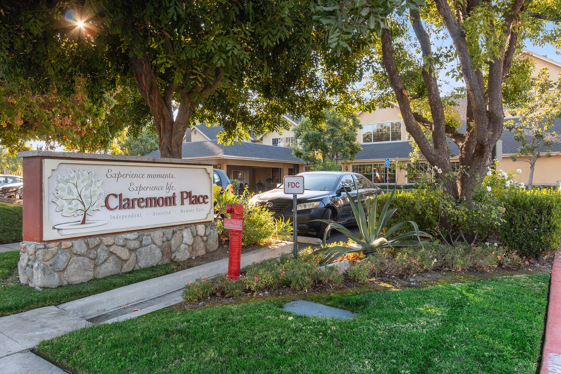 Outdoor view of Claremont Place senior living facility sign with trees, plants, a fire department connection, and a parked black car in front of the building under a sunny sky.