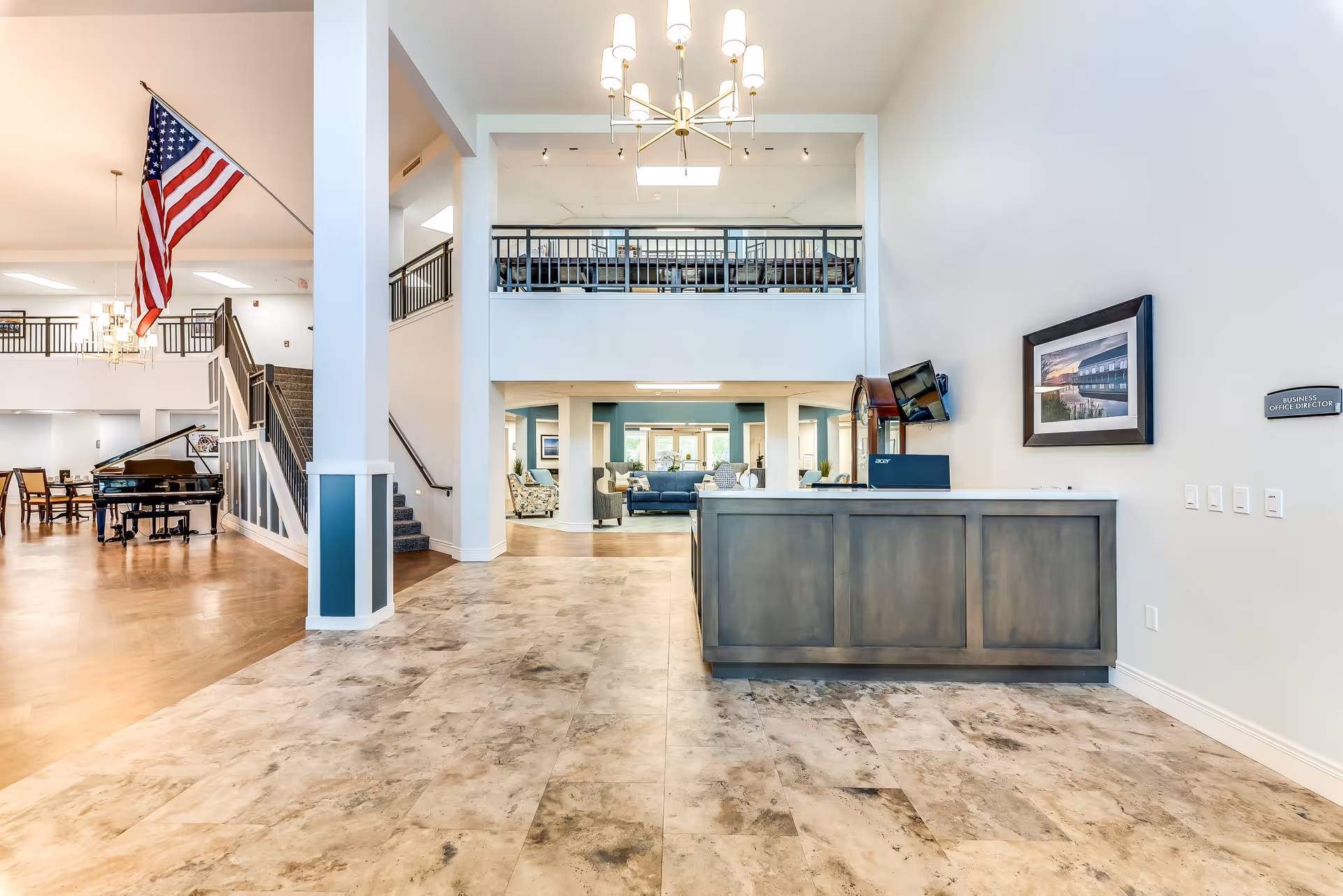 Bright and spacious senior living facility lobby with a reception desk on the right, a grand piano and dining area to the left, and a staircase leading to an upper balcony. The space features high ceilings, a chandelier, an American flag, and comfortable seating areas in the background.
