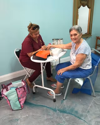 A woman in a maroon uniform providing a manicure to an elderly woman seated on a blue chair in a room with light blue walls and a large mirror on the wall.