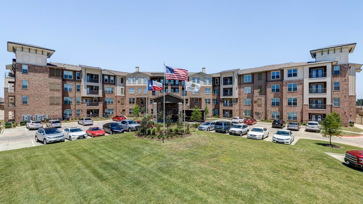 Four-story brick senior living building with a flagpole display, parked cars, and a large mowed lawn under a clear sky.