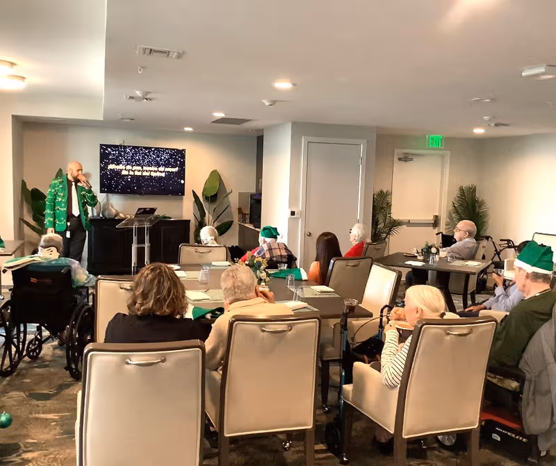 A group of elderly people seated at tables in a common room, watching a man in a green festive jacket speaking or singing in front of a TV screen. Some attendees are wearing green elf hats, and the room is decorated with plants and has a neutral color scheme.