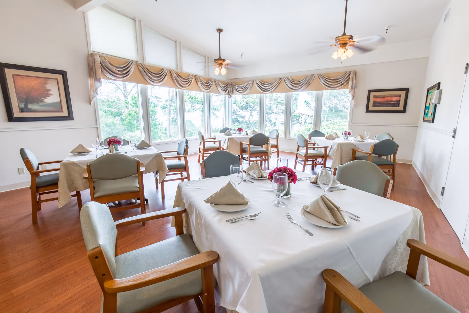 A bright dining room with several tables covered in white tablecloths, each set with folded napkins, glasses, and silverware. The room has large windows with a view of trees outside, wooden chairs with green cushions, ceiling fans with lights, and framed artwork on the walls.