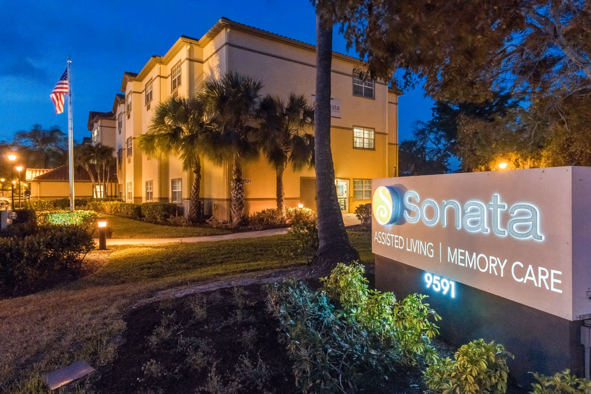 Exterior view of Sonata Boca Raton assisted living and memory care facility at dusk, showing a well-lit building, palm trees, an American flag, and a large illuminated sign with the facility's name and address.