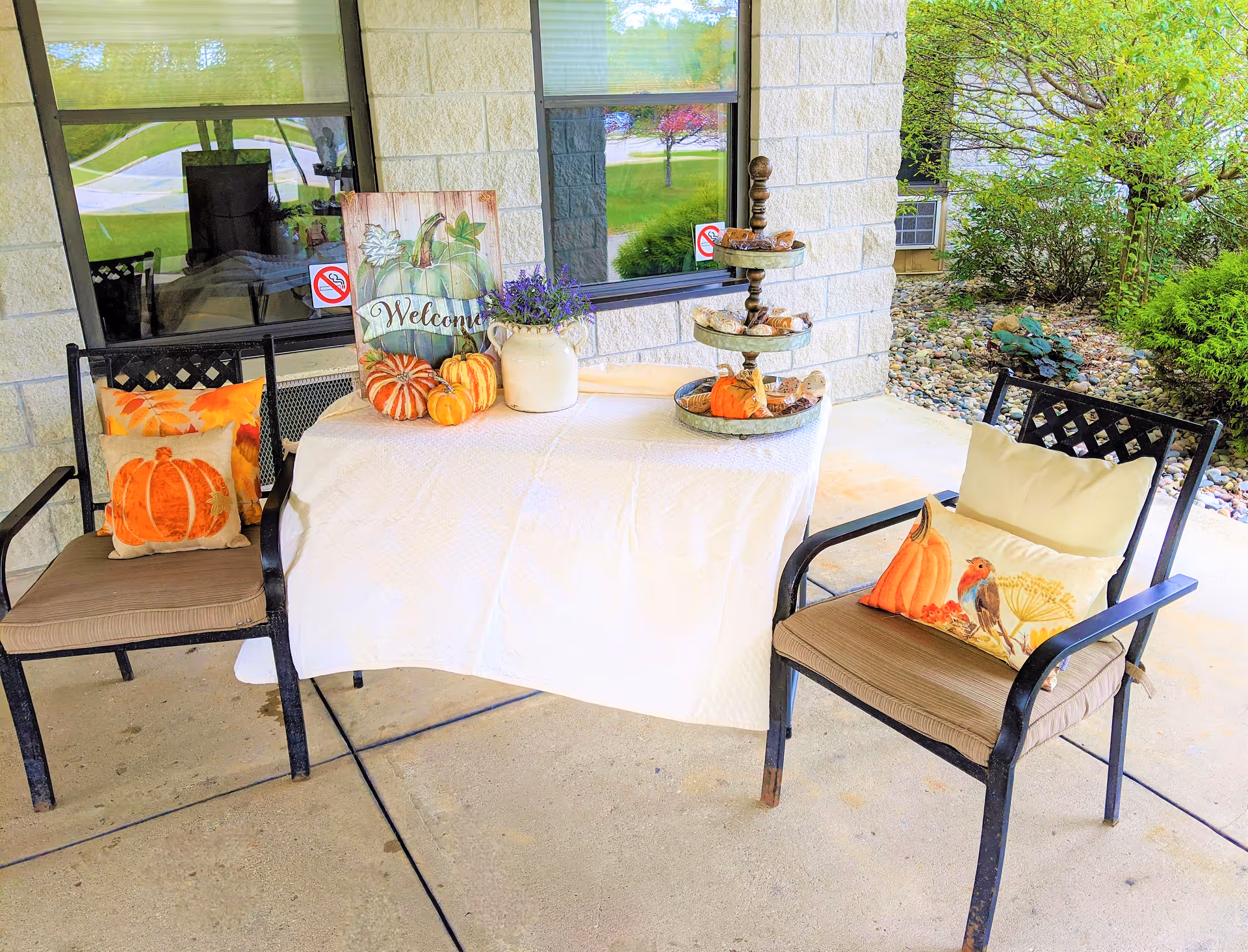 Outdoor patio area with two black metal chairs with brown cushions and decorative fall-themed pillows featuring pumpkins and birds. A small table covered with a white cloth holds a welcome sign with pumpkins, a vase with purple flowers, and a three-tiered tray with decorative items. The setting is next to a building with windows and surrounded by greenery and rocks.