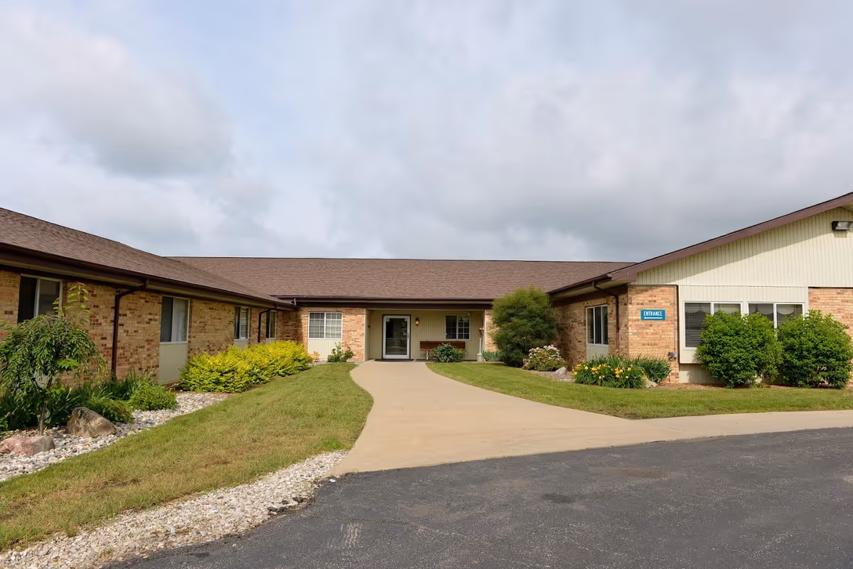 Exterior view of a single-story brick building with a brown roof and a paved walkway leading to the entrance. The building is surrounded by green grass, bushes, and small landscaped areas with rocks. A blue sign near the entrance reads 'ENTRANCE'. The sky is partly cloudy.