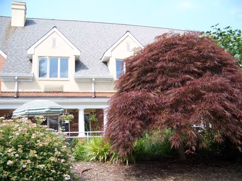 Exterior view of a residential building with beige siding and a gray shingled roof. The building has two dormer windows and a porch with white columns. In the foreground, there is a landscaped garden with a large reddish-brown tree, green shrubs, and flowering plants. A striped green and white umbrella is visible on the porch.