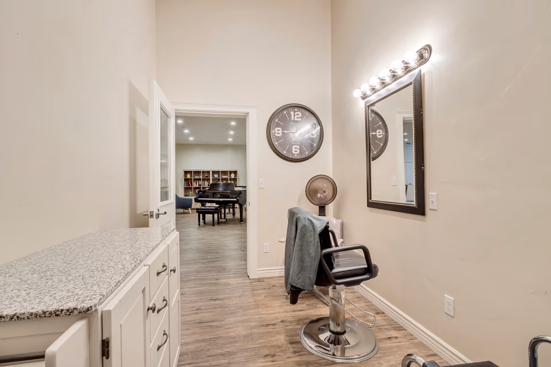 A small salon area with a black salon chair facing a wall-mounted mirror with lights above it. A round wall clock is mounted on the wall next to the mirror. A countertop with white cabinets is on the left side. Through an open door, a room with a grand piano and bookshelves is visible.
