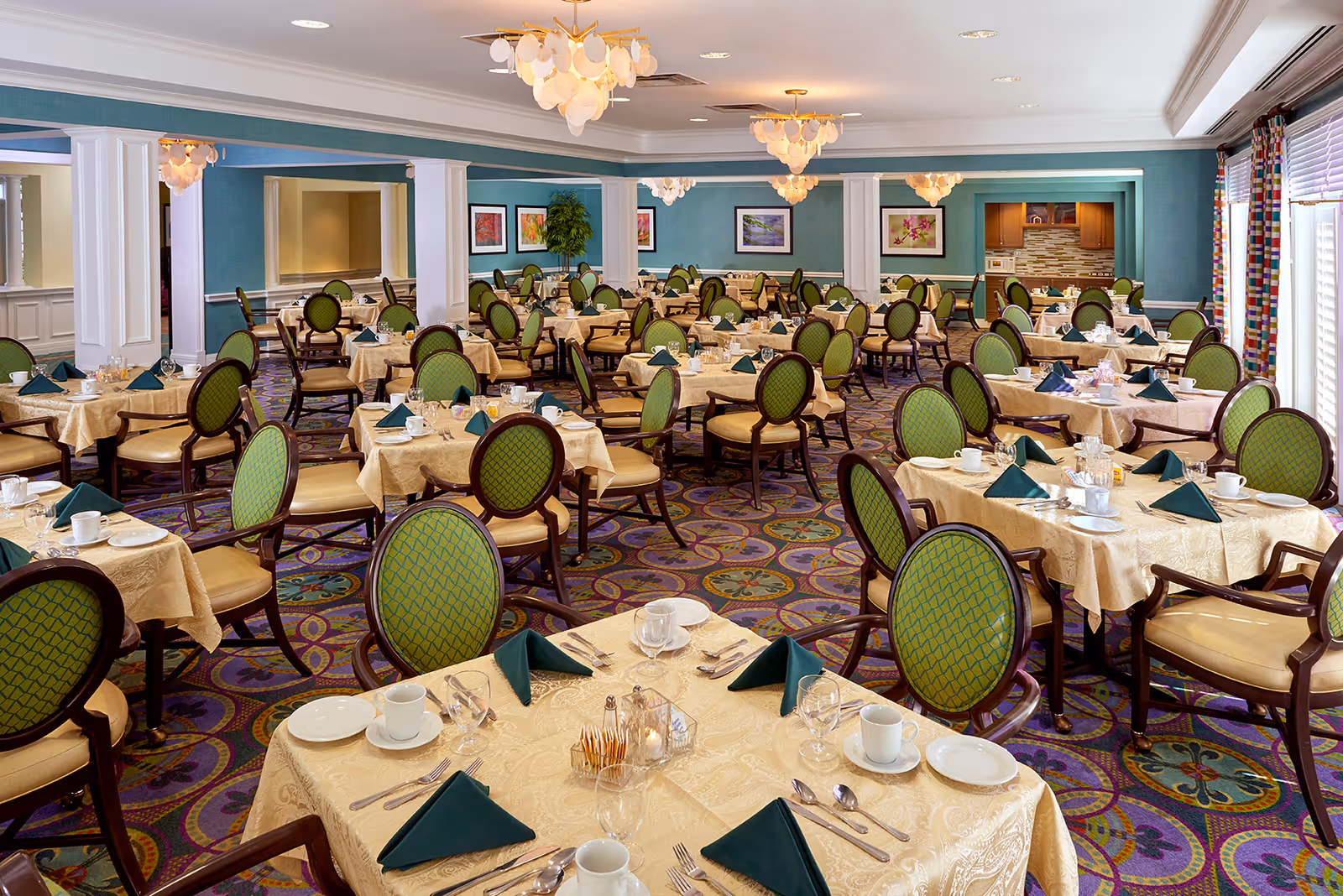 A spacious dining room with multiple tables covered in beige tablecloths, each set with white plates, cups, silverware, and green folded napkins. The chairs have green cushioned backs and seats with wooden frames. The room features patterned carpet, teal walls with white trim, framed artwork, and large windows with colorful curtains allowing natural light to enter. Elegant chandeliers hang from the ceiling.