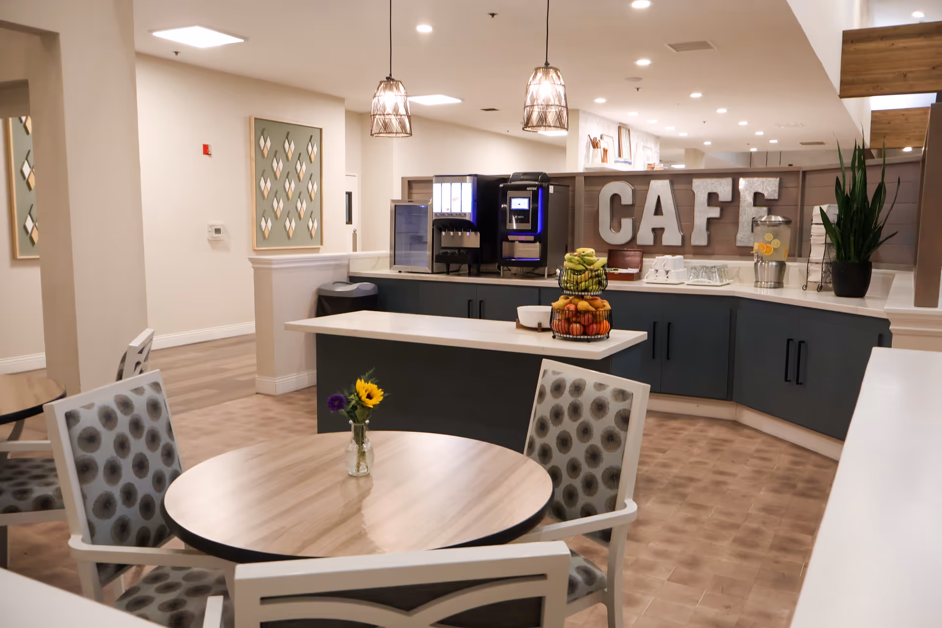 Interior view of a senior living facility cafe area with a round wooden table and four patterned chairs. On the table is a small vase with a sunflower and other flowers. In the background, there is a counter with a coffee machine, a beverage dispenser with lemon water, a tiered fruit basket, and large letters spelling 'CAFE' on the wall behind the counter. The space is well-lit with pendant lights and recessed ceiling lights.