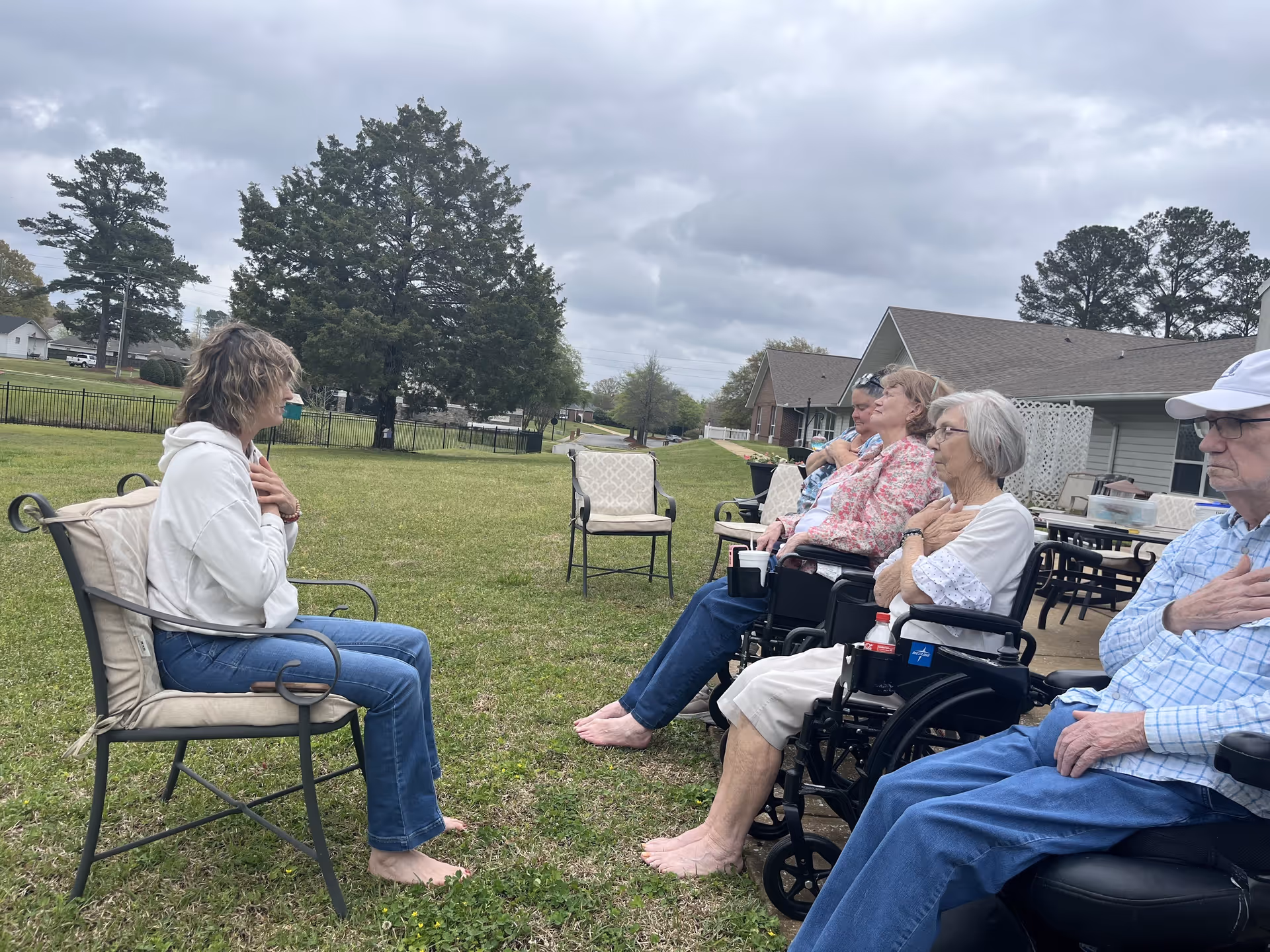 A group of elderly people sitting outside on chairs and wheelchairs on a grassy area, facing a woman seated on a cushioned chair. The woman and the elderly individuals have their hands placed on their chests, appearing to engage in a calming or meditative activity. The background shows trees, a fence, and residential buildings under a cloudy sky.